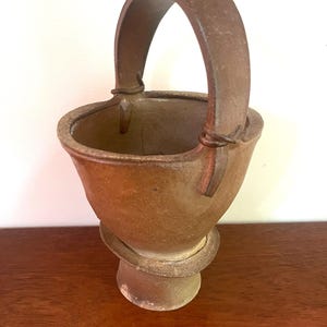 May include: A brown ceramic basket with a curved handle. The basket has a wide opening and a rounded base, sitting on a pedestal. The handle is attached with wire. The basket is on a wooden surface.