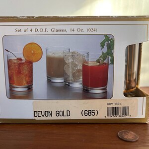 May include: Set of four 14 ounce Devon Gold glasses. The glasses are clear and have a round shape. The box has the text "Set of 4 D.O.F. Glasses, 14 Oz. (024)" and "DEVON GOLD (685)" printed on it.