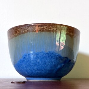 May include: A ceramic bowl featuring a smooth, glossy glaze that transitions from a deep, vibrant blue at the base to a lighter, muted blue in the centre, topped with a textured, speckled brown rim. A small coin is placed next to the bowl for scale.