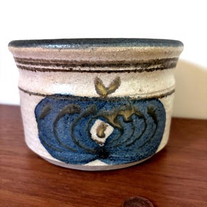 May include: A small, round ceramic pot with a blue floral design. The pot has a cream-colored base with brown and black stripes near the rim. A penny is next to the pot for scale. The pot is on a wooden surface.