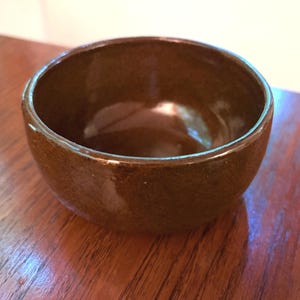 May include: A small, round, deep bowl with a smooth, glossy finish in a solid dark brown color. The bowl features a simple, curved silhouette with a wide opening and a consistent, reflective glaze covering the entire exterior and interior surface.