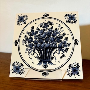 May include: A square ceramic tile with a hand-painted design of a basket overflowing with fruit and foliage in shades of blue. The tile is framed by a circular border and corner floral accents. The tile is displayed on a clear acrylic stand.