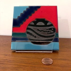 May include: A ceramic tile with a Southwestern design. The tile features a black and gray vase with a wavy pattern, set against a red and blue background with a stepped design. The tile is displayed in a clear acrylic stand.