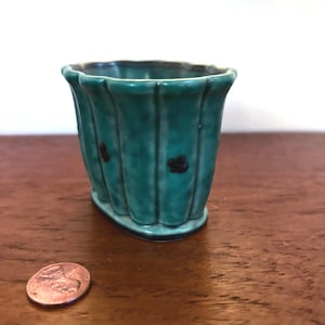 May include: A small, teal-colored ceramic vase with a fluted design and dark brown accents. The vase has a rectangular shape with rounded edges and small, dark brown floral details. A penny is next to the vase.