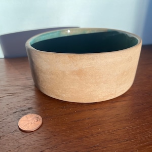 May include: A small, round, ceramic bowl with a light brown exterior and a dark green interior. The bowl is sitting on a wooden surface.