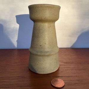 Puede incluir: Un jarrón de cerámica beige con una textura moteada. El jarrón tiene un cuerpo cilíndrico con una parte superior e inferior ensanchadas. Una moneda está al lado del jarrón para comparar el tamaño. El jarrón está sobre una superficie de madera.