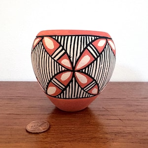 May include: A small, round, hand-painted ceramic bowl. The bowl features a coral-colored base and rim, with a black and white geometric pattern. The design includes vertical stripes and petal-like shapes. A penny is next to the bowl.