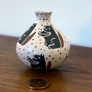 May include: A small, hand-painted ceramic vase with a rounded body and narrow neck. The off-white vase features black hippo illustrations and brown accents, with small black and brown dots. A penny is next to the vase.