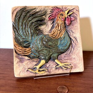 Può includere: Piastrella in ceramica quadrata raffigurante un gallo in rilievo. Il gallo è dipinto con colori rosso, blu, verde e marrone. La piastrella è esposta su un supporto trasparente. Una moneta è visibile accanto al supporto.
