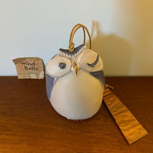 May include: A ceramic owl wind chime with blue and white paint, hanging from a gold cord. The owl has one eye closed and the other open. The tag attached to the owl reads "Wind Bells".