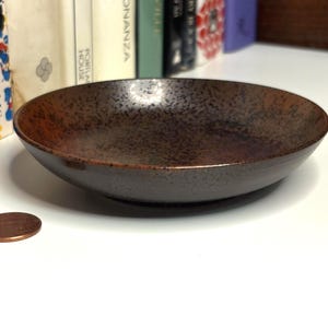 Puede incluir: Un cuenco circular poco profundo con un acabado de esmalte moteado de color marrón oscuro. El cuenco presenta un borde curvo y liso y un interior ancho y abierto. Una moneda de color cobre está colocada junto al cuenco para dar escala al objeto.