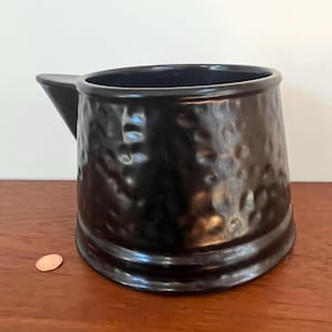 May include: A black ceramic pitcher with a hammered texture. The pitcher has a wide mouth and a spout.