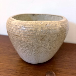 Op de afbeelding: Een kleine, handgemaakte, beige keramische kom met een afgeronde, rechthoekige opening. De kom heeft een getextureerd oppervlak met horizontale lijnen en een licht taps toelopende vorm. Een munt ligt naast de kom.