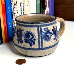 May include: A speckled beige ceramic mug with a rounded body and a sturdy handle. The mug features a hand-painted blue floral pattern around the centre, bordered by horizontal blue lines. A copper-coloured penny sits on the surface nearby.