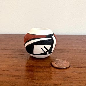 May include: A small, round ceramic vase with a white rim, featuring a geometric design in black, white, and reddish-brown. The vase is approximately 2 inches in diameter. A penny is next to the vase for size comparison.