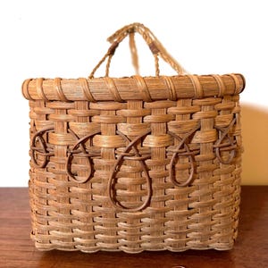 May include: A rectangular woven basket with a braided handle. The basket is made of light brown and tan woven material. Decorative teardrop shapes are woven into the front of the basket. The basket is sitting on a wooden surface.