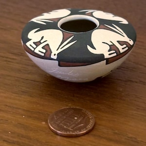 May include: A small, round, hand-painted ceramic bowl with a dark green rim and a cream-colored body. The rim features white rabbit designs. The bowl is approximately 3 inches in diameter. A penny is next to the bowl for size comparison.