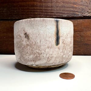 May include: A cylindrical ceramic bowl with a textured, matte off-white and light brown mottled glaze, featuring a single vertical dark charcoal drip mark on the side, placed next to a copper-colored United States one-cent coin.