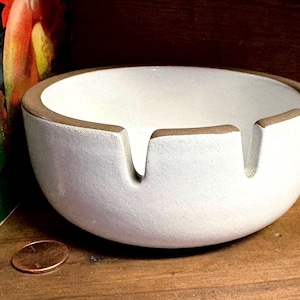May include: A round, white ceramic ashtray with a smooth, matte finish and a contrasting brown rim. The bowl features three evenly spaced notches along the top edge for resting items. The design is simple, circular, and functional in its construction.