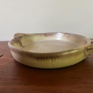 May include: A small, round ceramic dish with two handles. The dish has a light beige interior and a darker brown rim, with a glossy finish. The dish is on a wooden surface.