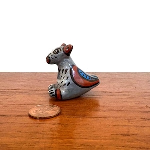 May include: A small, gray ceramic animal figurine with red and blue accents. The figurine is in a seated position with its paws in front. A penny is next to the figurine. The figurine has black spots and a red nose.