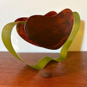May include: A decorative heart-shaped object with a dark brown, marbled appearance. The heart is supported by a looping, light green ribbon. A penny is placed on the wooden surface below the heart.