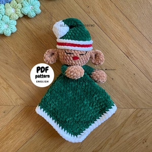 May include: A green and white crocheted elf doll with a red and white striped hat. The doll is lying on a green and white crocheted blanket. The blanket has a white border. The doll is holding its hands together in a praying position. The doll is on a wooden surface. The text "PDF pattern ENGLISH" is on a white circle in the lower left corner of the image.
