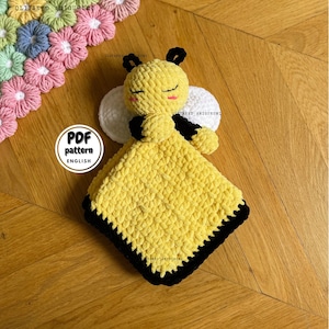 Puede incluir: Una manta de bebé de ganchillo en forma de abeja con un cuerpo amarillo, rayas negras y alas blancas. La manta tiene un borde negro y está sobre una superficie de madera. El texto "PDF pattern ENGLISH" es visible en una etiqueta blanca.