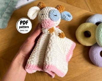 Op de afbeelding: Een gehaakte koe-vormige veiligheidsdeken met een roze neus, blauwe ogen en bruine oren. De deken is wit met een roze rand en wordt in een hand gehouden. De tekst "PDF pattern ENG/SK" is zichtbaar in de afbeelding.
