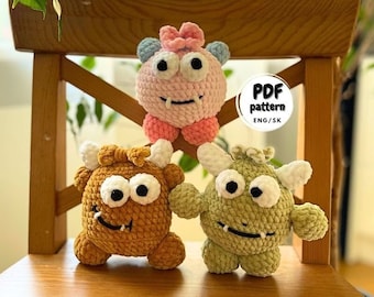 May include: Three crocheted plush toys in the shape of monsters. The monsters are pink, brown, and green. The pink monster has a bow on its head. The brown monster has horns. The green monster has a toothy grin. The toys are sitting on a wooden chair. PDF pattern ENG/SK