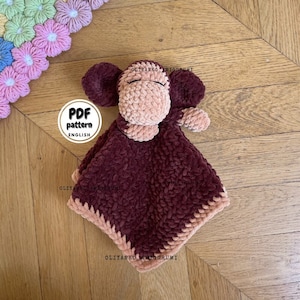 May include: A crocheted monkey amigurumi toy with a brown and pink color scheme. The toy is lying on a wooden surface. The text "PDF pattern ENGLISH" is visible on a white tag attached to the toy.