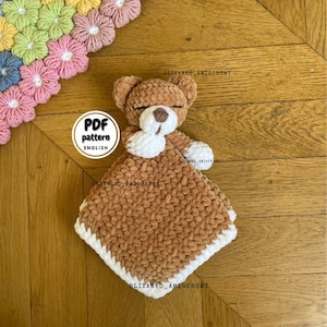 May include: A brown crocheted blanket with a white border and a small brown crocheted bear with white accents lying on top. The blanket and bear are on a wooden surface. The text "PDF pattern ENGLISH" is visible on a white circle.