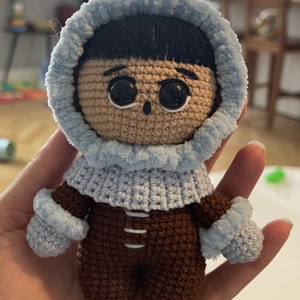 Puede incluir: Un juguete de peluche tejido a crochet que lleva una chaqueta con capucha azul y pantalones marrones. El juguete tiene el pelo negro, los ojos marrones y una sonrisa cosida.