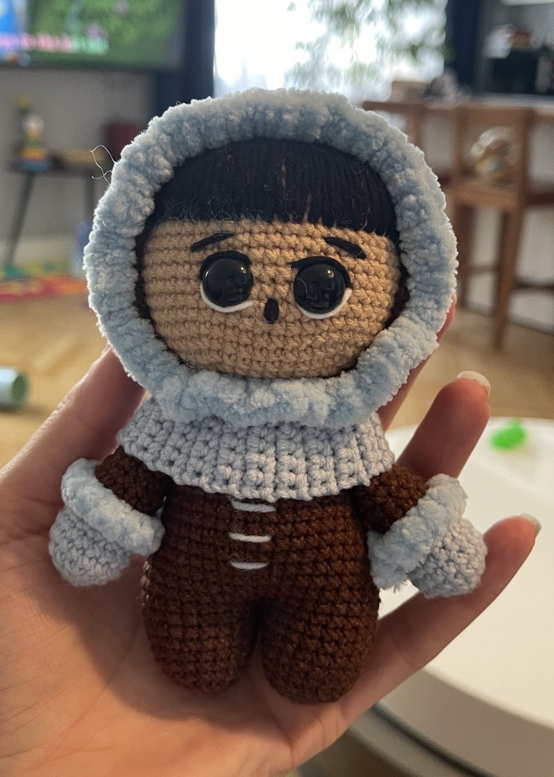 Puede incluir: Un juguete de peluche tejido a crochet que lleva una chaqueta con capucha azul y pantalones marrones. El juguete tiene el pelo negro, los ojos marrones y una sonrisa cosida.