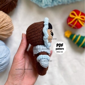 Puede incluir: Una mu&ntilde;eca amigurumi de crochet marr&oacute;n que lleva un gorro y un abrigo de crochet azul. La mu&ntilde;eca est&aacute; siendo sostenida en una mano. El texto "PDF pattern ENG/SK" es visible en la imagen.