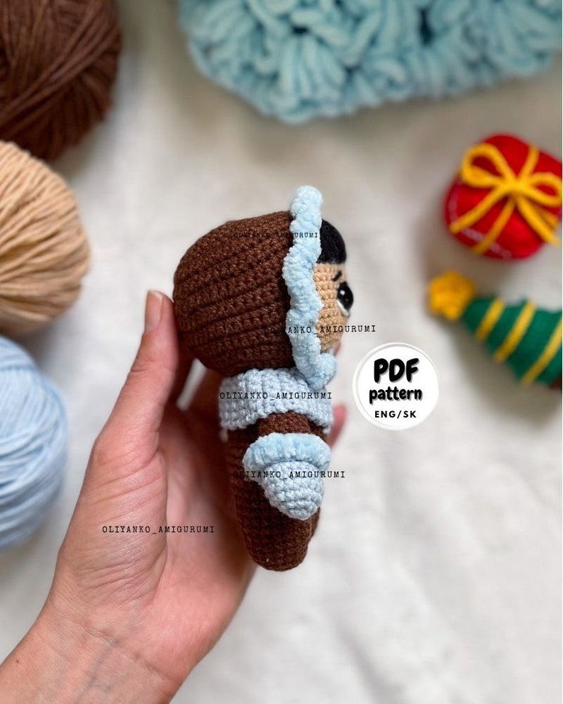 Puede incluir: Una mu&ntilde;eca amigurumi de crochet marr&oacute;n que lleva un gorro y un abrigo de crochet azul. La mu&ntilde;eca est&aacute; siendo sostenida en una mano. El texto "PDF pattern ENG/SK" es visible en la imagen.
