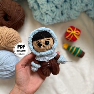 Puede incluir: Mu&ntilde;eca amigurumi de crochet con una chaqueta con capucha azul y pantalones marrones. La mu&ntilde;eca tiene el pelo negro y los ojos marrones. La mu&ntilde;eca sostiene una peque&ntilde;a caja de regalo y un adorno de &aacute;rbol de Navidad. El texto "PDF pattern ENG/SK" es visible en la imagen.