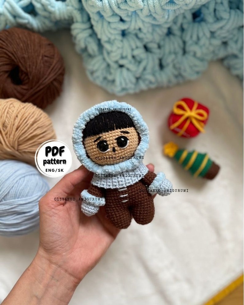 Puede incluir: Mu&ntilde;eca amigurumi de crochet con una chaqueta con capucha azul y pantalones marrones. La mu&ntilde;eca tiene el pelo negro y los ojos marrones. La mu&ntilde;eca sostiene una peque&ntilde;a caja de regalo y un adorno de &aacute;rbol de Navidad. El texto "PDF pattern ENG/SK" es visible en la imagen.