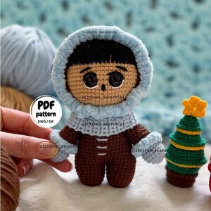 Puede incluir: Mu&ntilde;eca amigurumi de crochet con un atuendo de invierno con capucha azul y pantalones marrones. La mu&ntilde;eca tiene cabello negro y ojos marrones. La mu&ntilde;eca est&aacute; de pie junto a un peque&ntilde;o &aacute;rbol de Navidad de crochet con una estrella amarilla en la parte superior.
