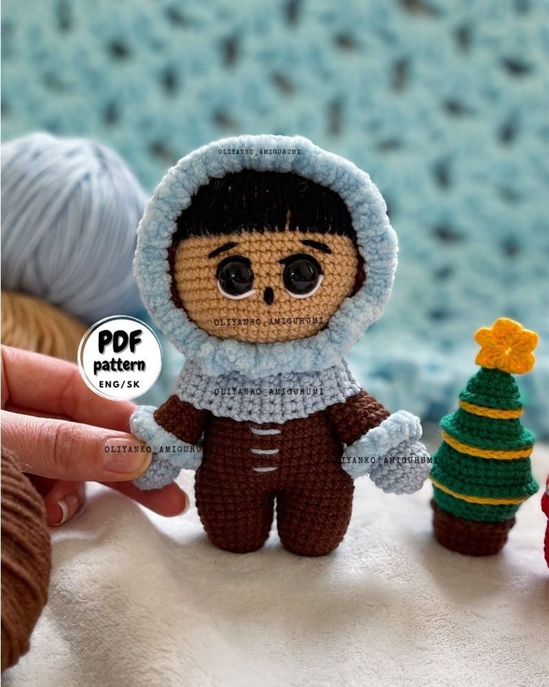 Puede incluir: Mu&ntilde;eca amigurumi de crochet con un atuendo de invierno con capucha azul y pantalones marrones. La mu&ntilde;eca tiene cabello negro y ojos marrones. La mu&ntilde;eca est&aacute; de pie junto a un peque&ntilde;o &aacute;rbol de Navidad de crochet con una estrella amarilla en la parte superior.