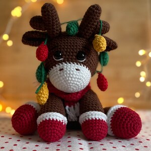 Crochet Reindeer, dark brown in color with white muzzle, he is wearing red and white Christmas shoes on all 4 legs, he has red and white scarfs around his neck and colorful Christmassy lights hanging on his antlers. He is 13sc tall then sitting.