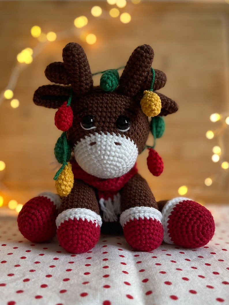 Crochet Reindeer, dark brown in color with white muzzle, he is wearing red and white Christmas shoes on all 4 legs, he has red and white scarfs around his neck and colorful Christmassy lights hanging on his antlers. He is 13sc tall then sitting.