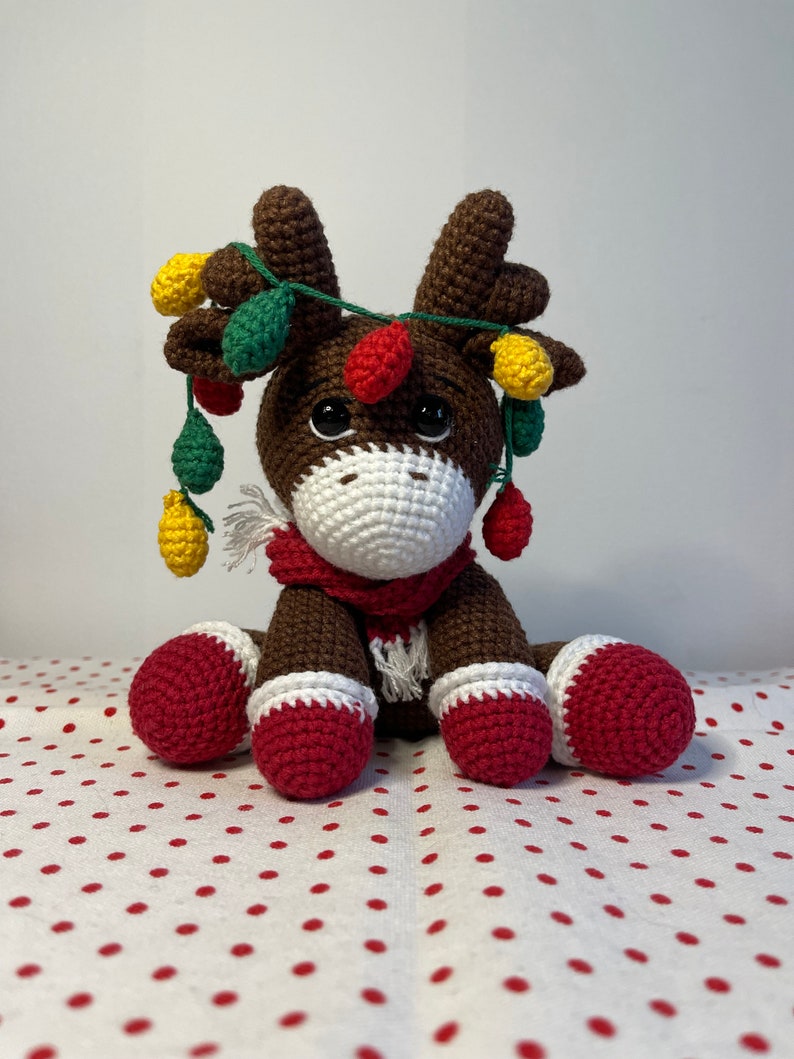 Crochet Reindeer, dark brown in color with white muzzle, he is wearing red and white Christmas shoes on all 4 legs, he has red and white scarfs around his neck and colorful Christmassy lights hanging on his antlers. He is 13sc tall then sitting.