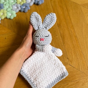 Puede incluir: Un conejito de crochet gris con una manta de crochet blanca adjunta. El conejito tiene una nariz rosa y los ojos cerrados.