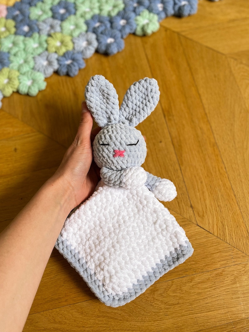Puede incluir: Un conejito de crochet gris con una manta de crochet blanca adjunta. El conejito tiene una nariz rosa y los ojos cerrados.