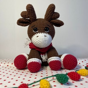 Crochet Reindeer, dark brown in color with white muzzle, he is wearing red and white Christmas shoes on all 4 legs, he has red and white scarfs around his neck and colorful Christmassy lights hanging on his antlers. He is 13sc tall then sitting.