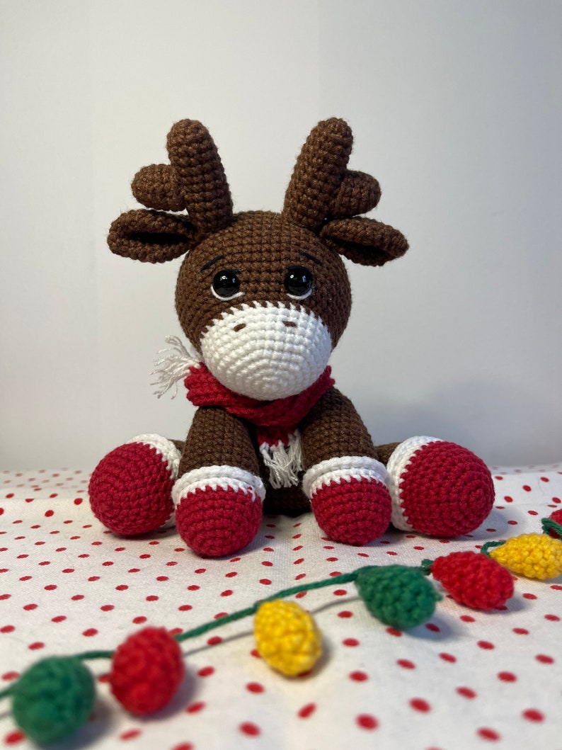 Crochet Reindeer, dark brown in color with white muzzle, he is wearing red and white Christmas shoes on all 4 legs, he has red and white scarfs around his neck and colorful Christmassy lights hanging on his antlers. He is 13sc tall then sitting.