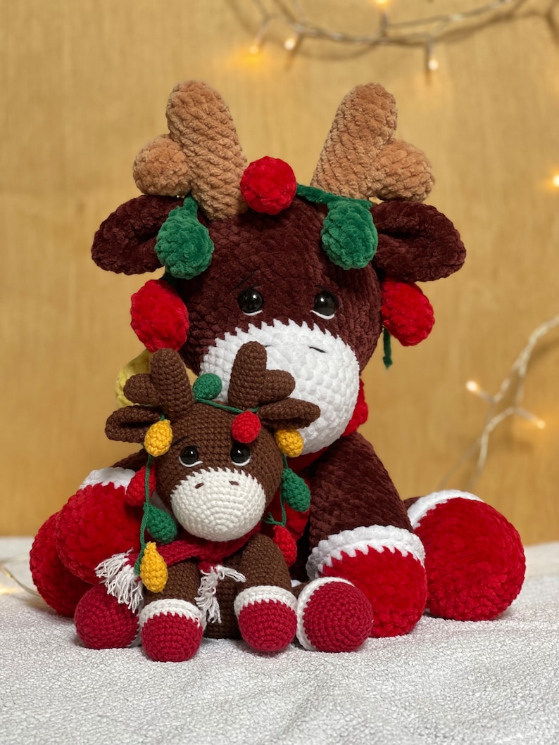 May include: Two crocheted reindeer ornaments, one large and one small, with red, green, and yellow lights. The reindeer are sitting on a white surface.