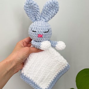Puede incluir: Un conejito de apego de crochet azul y blanco con la nariz rosa y los ojos cerrados. El conejito sostiene una manta de crochet blanca.