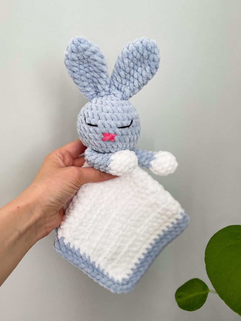 Puede incluir: Un conejito de apego de crochet azul y blanco con la nariz rosa y los ojos cerrados. El conejito sostiene una manta de crochet blanca.